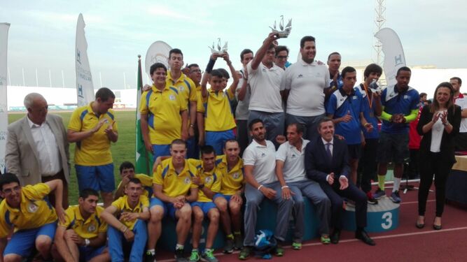 El equipo de La Línea, de blanco, en lo más alto del podio en el Estadio Iberoamericano de Huelva.