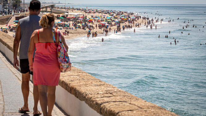 Dos personas pasean junto a la playa de Cádiz, repleta de bañistas.