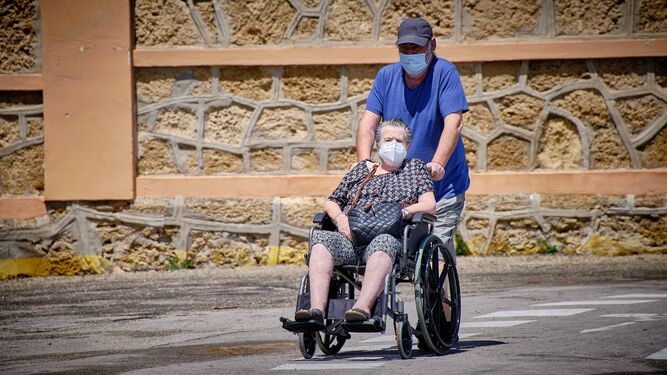 Una persona empuja a otra que va en silla de ruedas, ambas con mascarilla.